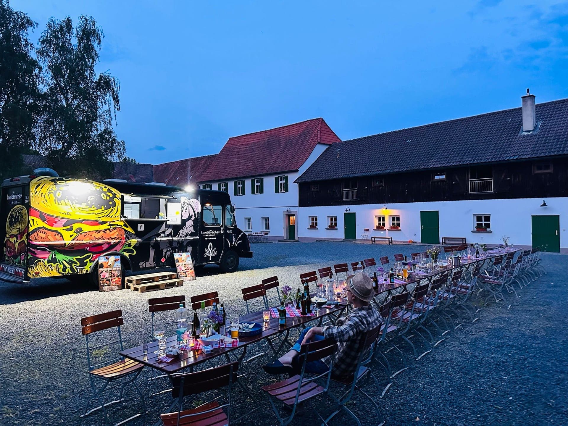 Lange gedeckte Tafel im Hof am Abend, daneben der beleuchtete Landstreicher-Truck