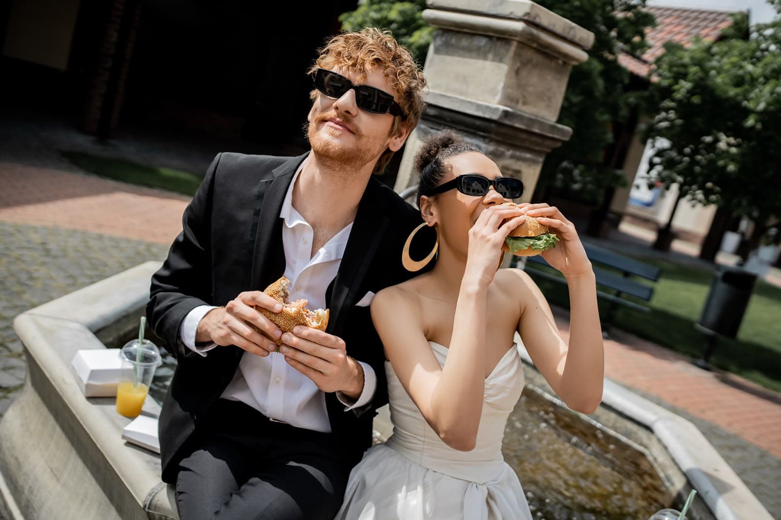 Brautpaar in Hochzeitsoutfit sitzt im Freien und isst Burger