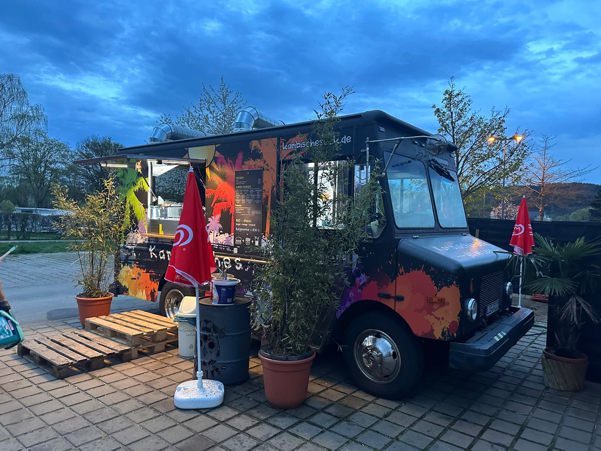 Der bunt bemalte Truck des Karibischen Ecks mit Palmenmotiven am Abend, davor Pflanzen und Paletten