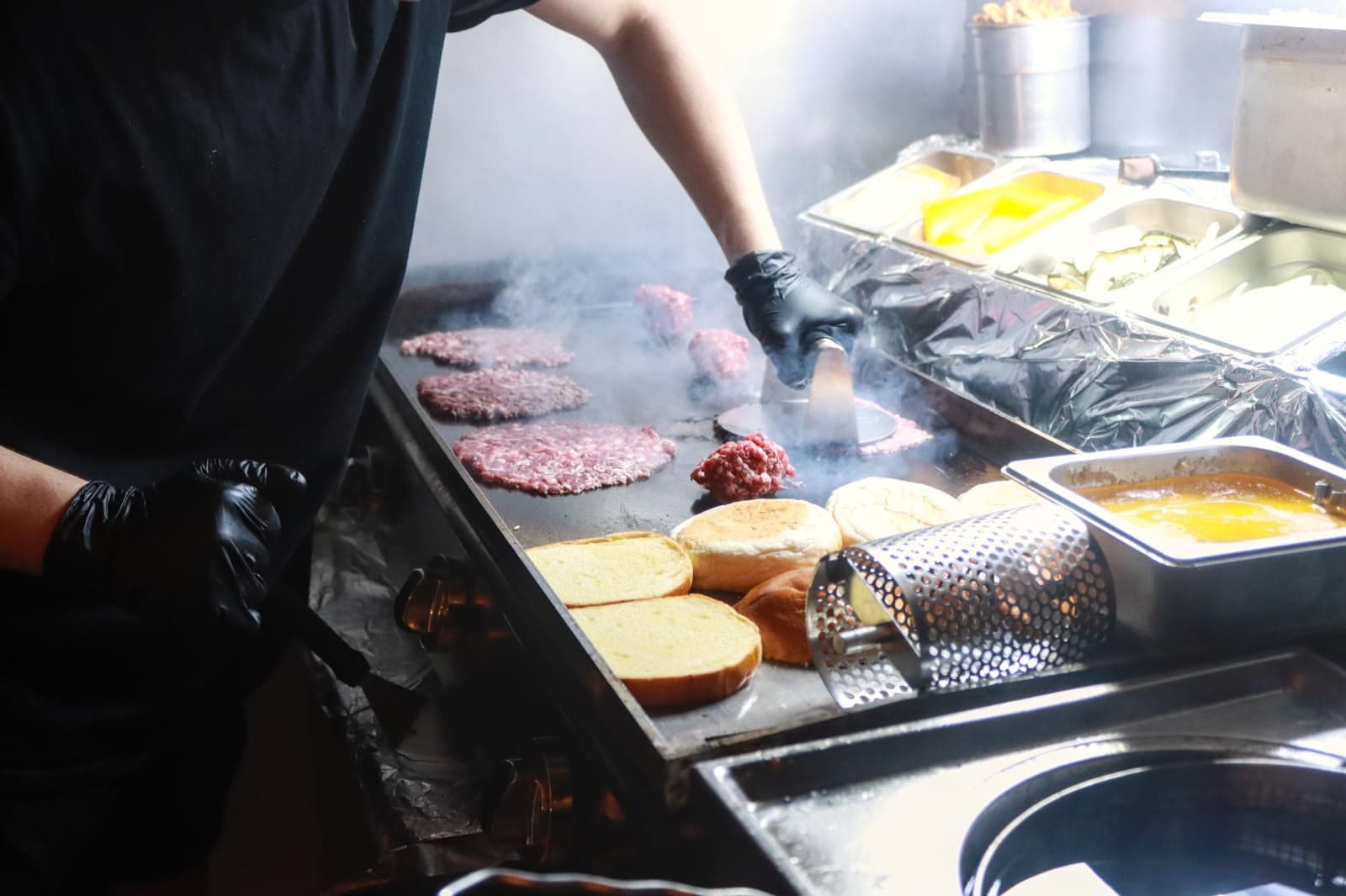 Koch drückt Rindfleisch-Patties auf der rauchenden Grillplatte, daneben Buns und Zutaten