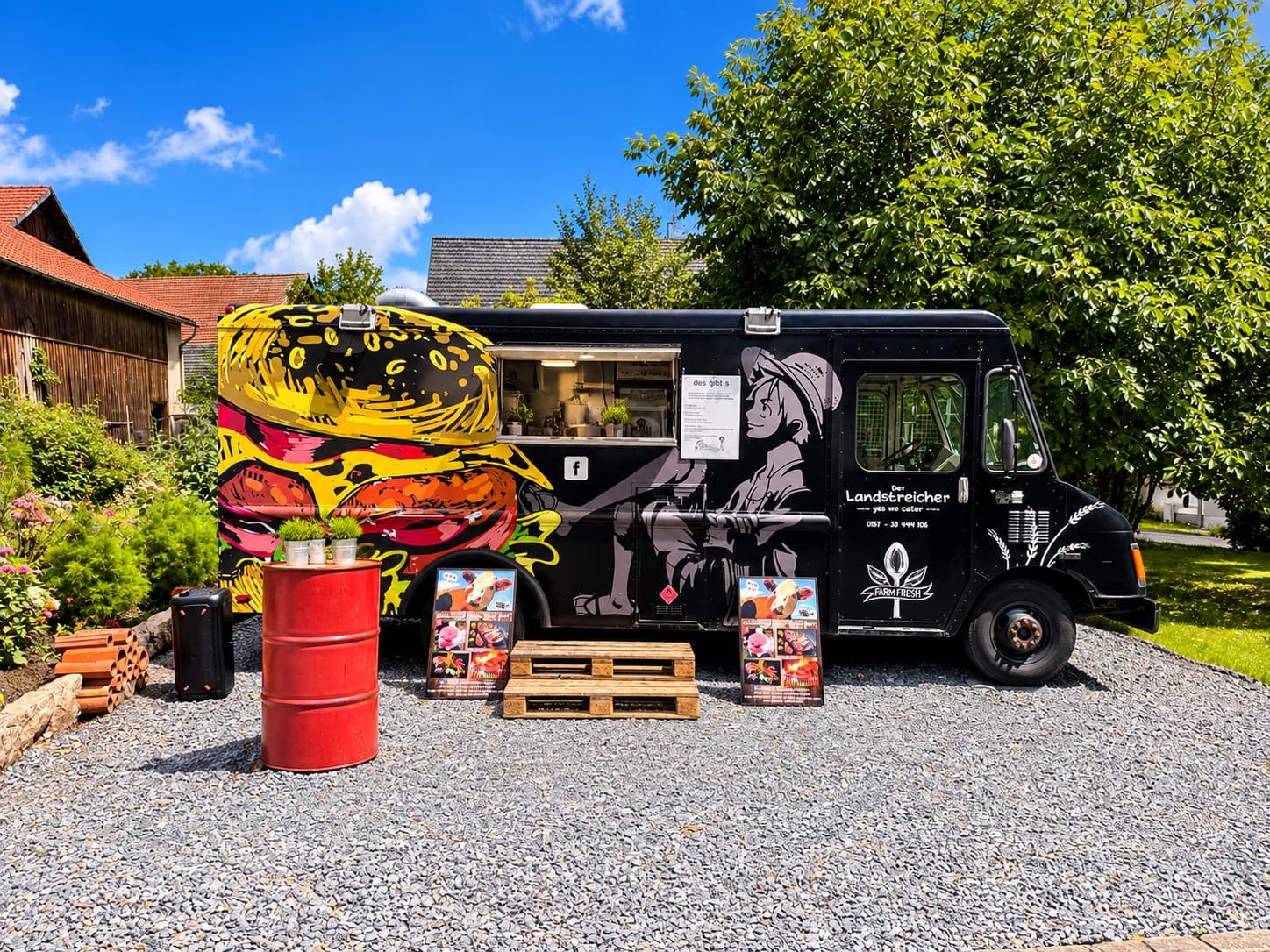 Der schwarze Landstreicher-Foodtruck mit Burger-Graffiti von der Seite, davor rotes Fass und Paletten auf Kies